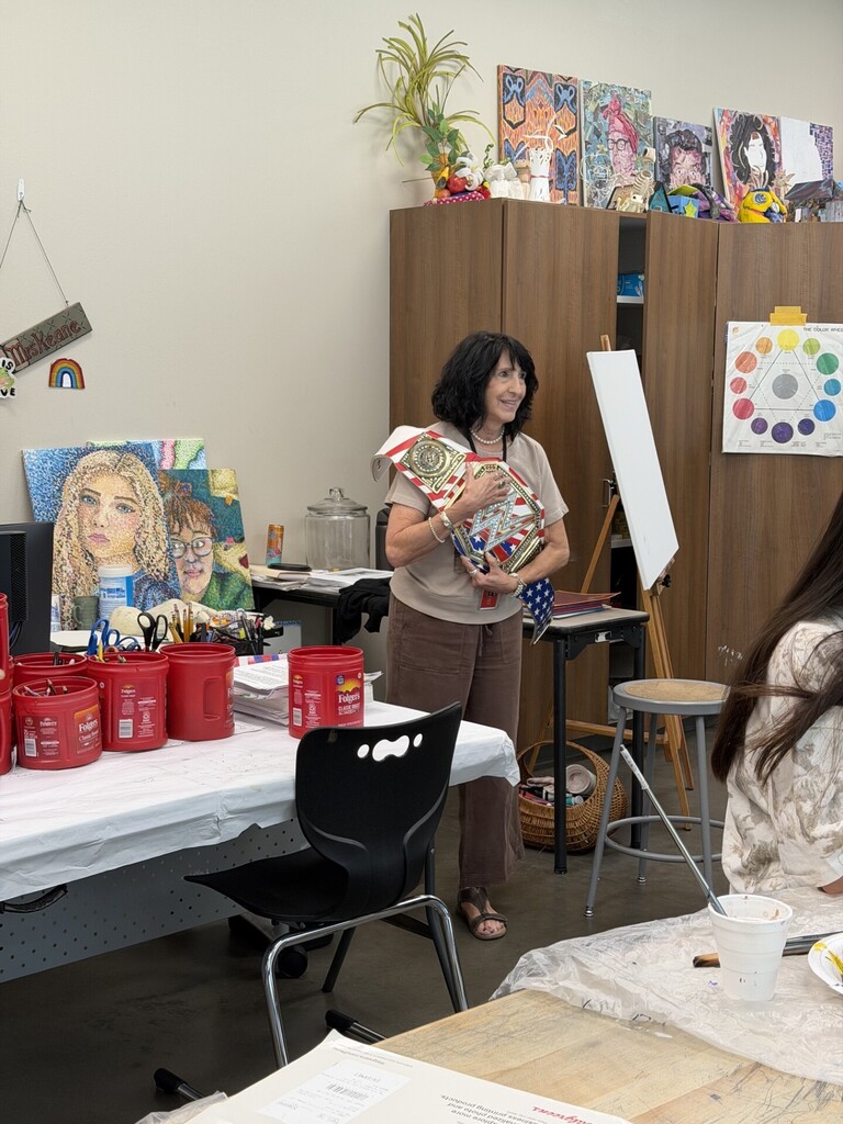 A room with art supplies and art pieces. A woman holds a craft. Tables and chairs are set up.