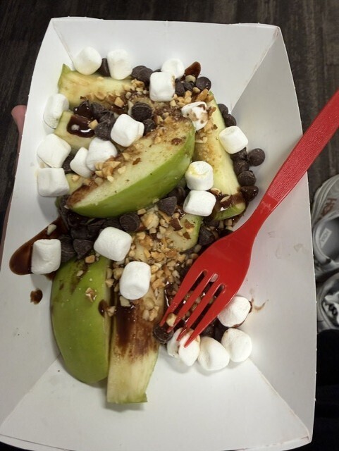 White box with dessert of green apple slices, caramel, chocolate chips, marshmallows, and nuts, topped with a red plastic fork.