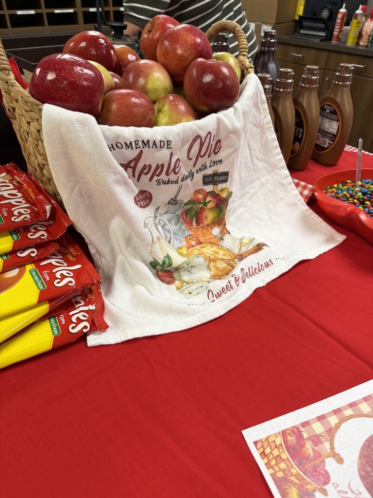 Basket of red apples on a table covered with a cloth reading "Homemade Apple Pie." Nearby are red tablecloth, candies, and bottles.