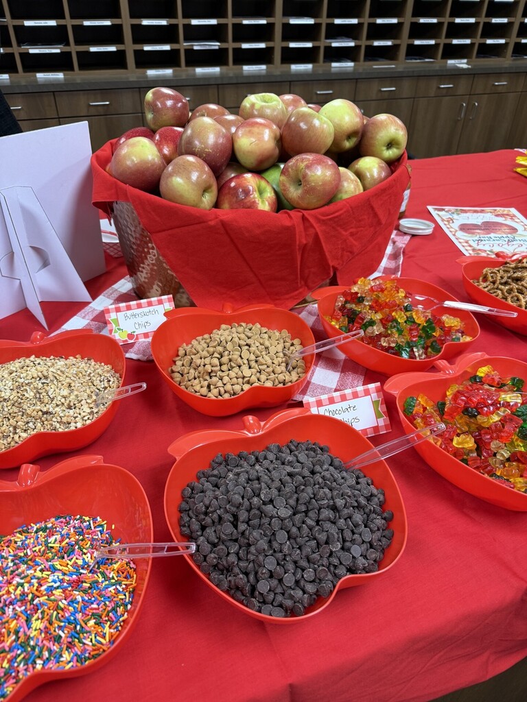 A table with a red tablecloth holds bowls of various treats in apple-shaped containers, including chips, beans, and candies.