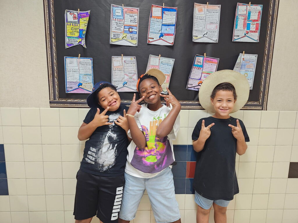 Three kids smile in front of a bulletin board with various papers pinned on it.