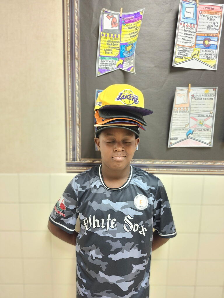 A child wears a camouflage shirt with "White Sox" and a yellow baseball cap. They stand in front of a bulletin board with colorful papers.