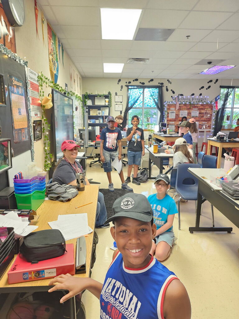 Classroom interior with several students seated at tables. A boy wearing a cap smiles with his hand raised.