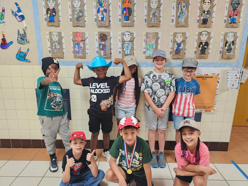 Nine children pose together in a classroom with hats and medals. A tiled floor and wall with art displays are behind them.