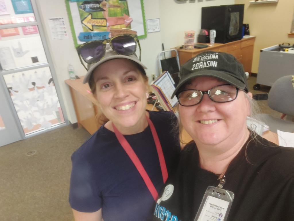 Two women in a room. One wears a hat, glasses, and a lanyard; the other has a hat and sunglasses.
