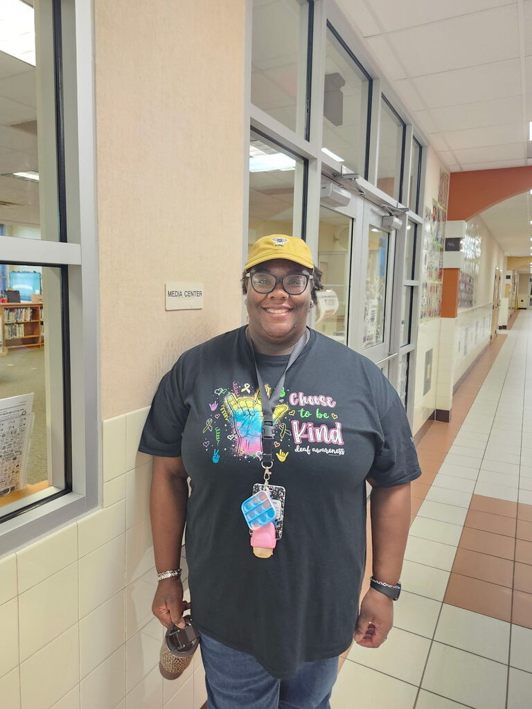 A person in a yellow hat and glasses stands in a school hallway with a "Choose to be Kind" shirt.