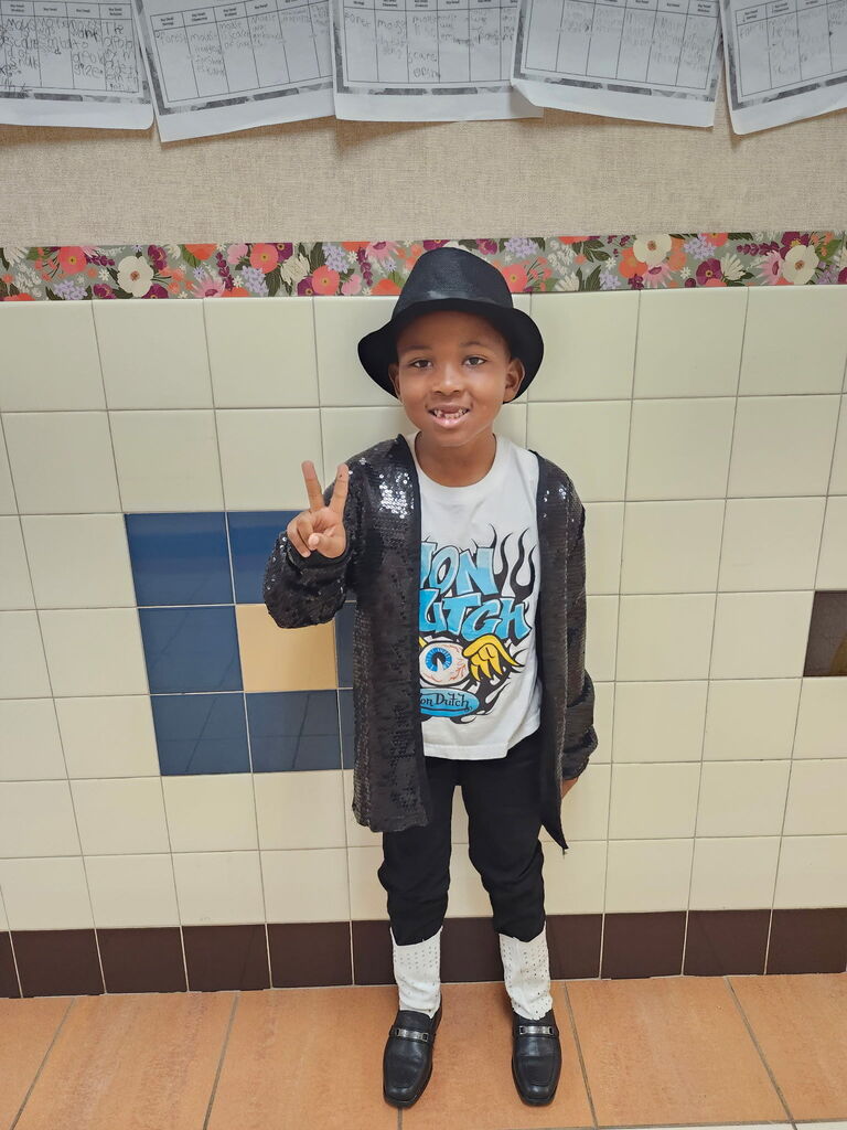 A child in a black hat, jacket, and black pants stands in a tiled corridor, making a peace sign.