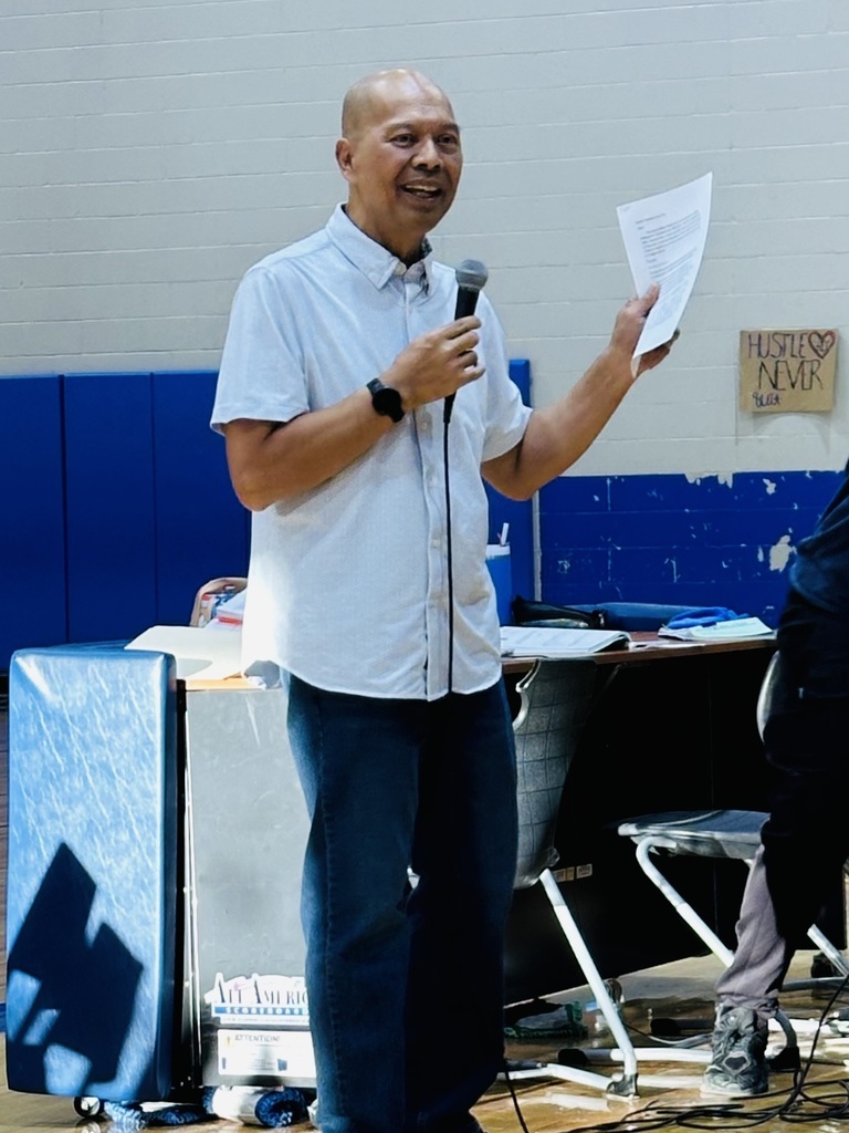 Person with a microphone in hand speaks at a gathering. Behind him, a desk holds papers and objects.