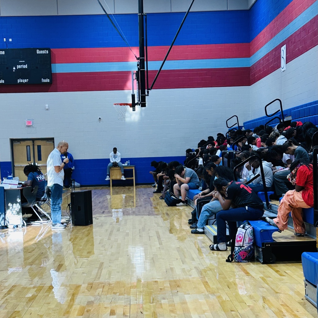 A basketball court with people seated on benches and a speaker. A man stands near a table with a microphone.