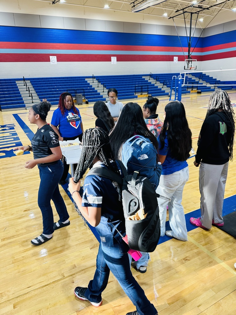 Several people, some with backpacks, converse in a gym with wooden flooring and blue and red walls.