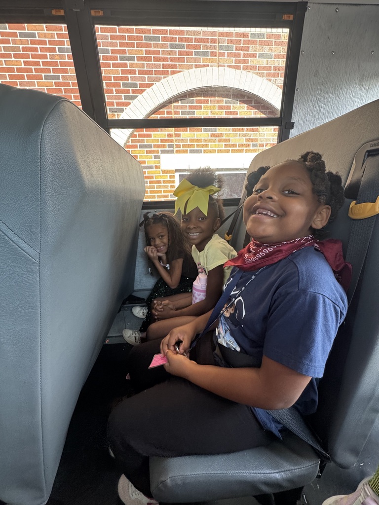 Three children seated inside a bus, one smiling. The bus has gray seats and a red brick building visible through the window.