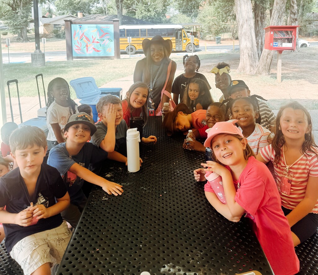Children seated around a table at a park, with a bus and trees in the background.