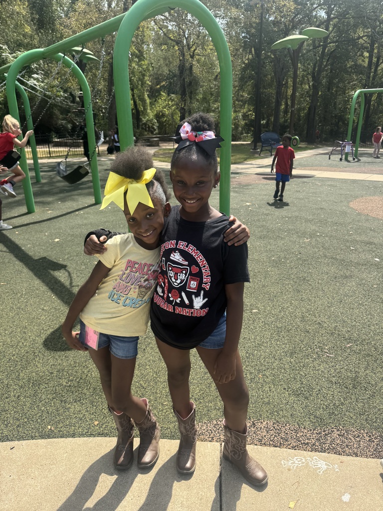 Two girls, one with a yellow bow, stand next to a playground with green metal equipment.