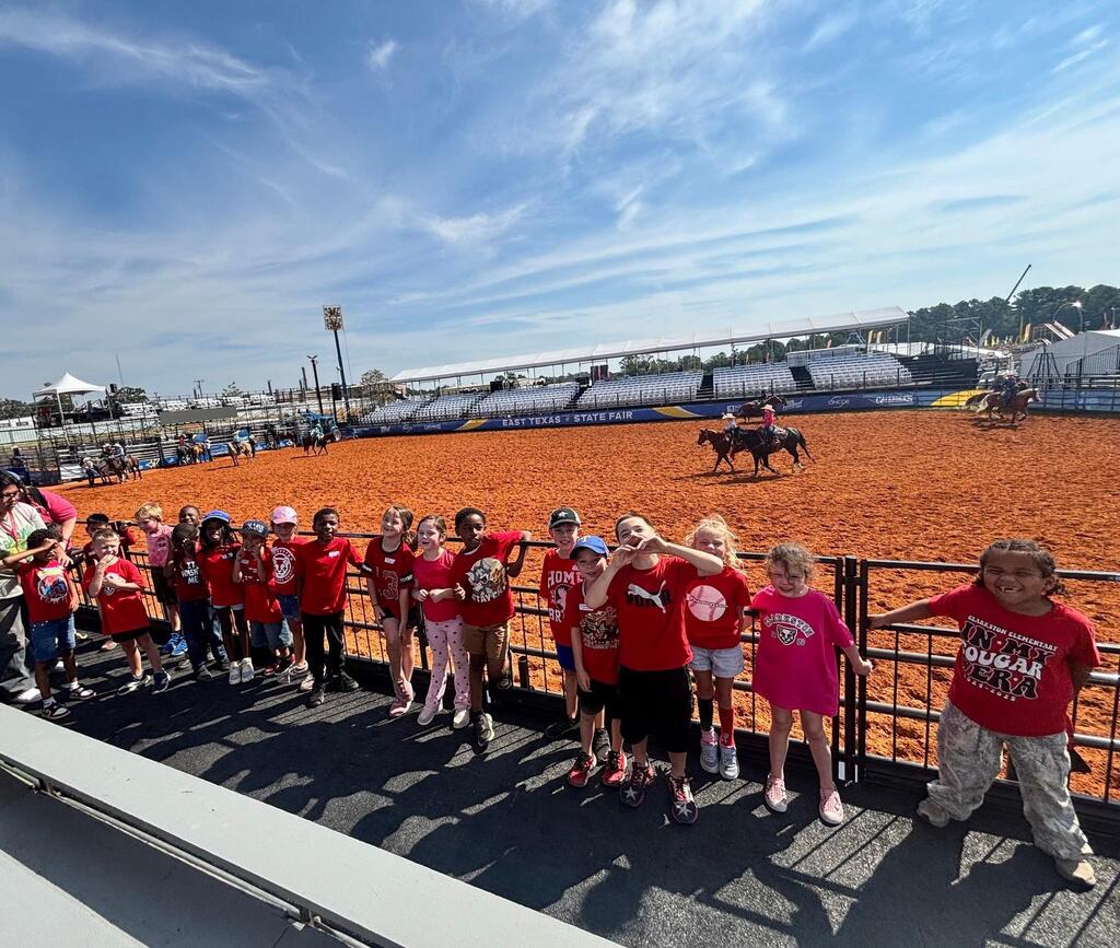 A group of Mrs. Fry's class in red shirts standing in front of a fence, watching a rodeo event.