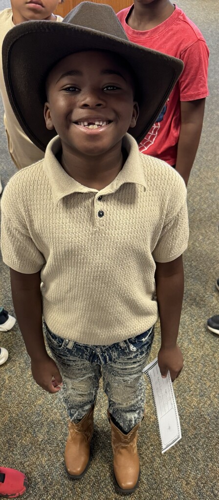 A young boy wearing a cowboy hat and boots smiles at the camera. He holds a piece of paper. Another child is in a red shirt behind him.