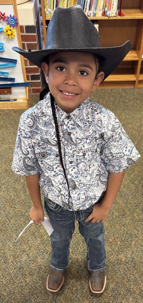 A young child in a cowboy hat and patterned shirt stands in a room with bookshelves in the background.