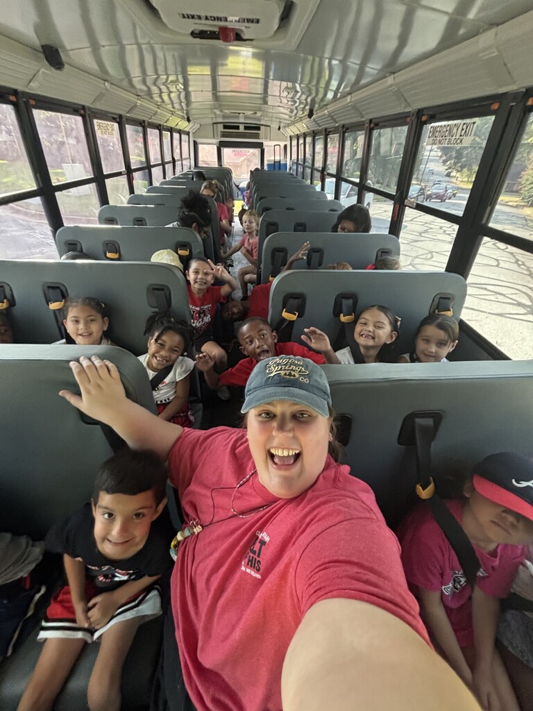 A bus with children seated, a woman in a pink shirt takes a selfie, children wearing hats and backpacks.