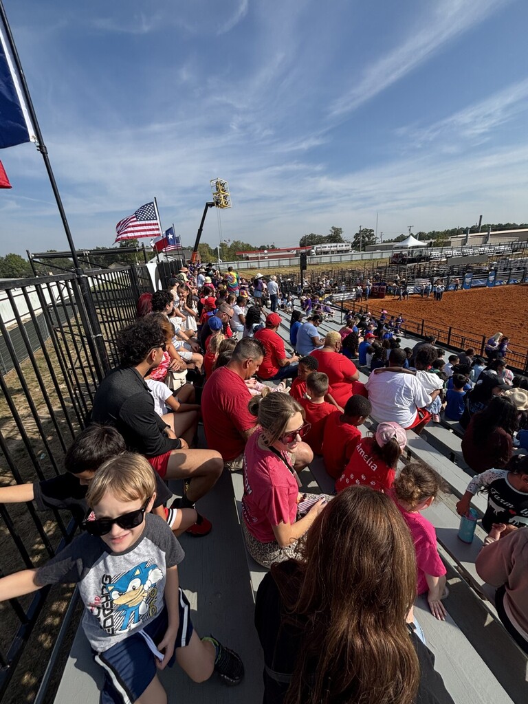 Spectators wearing red clothing watch from the bleachers at an outdoor sports venue with flags.