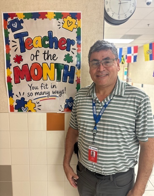 Man with ID card stands by a wall. A clock, colorful banners, and a banner reading "Teacher of the Month" are visible.