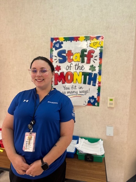 A woman in a blue shirt stands next to a "Staff of the Month" poster.