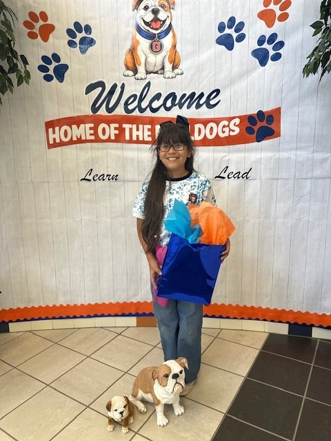 Girl with glasses holds a blue gift bag and stands in front of a banner reading "Welcome, Home of the Bulldogs".