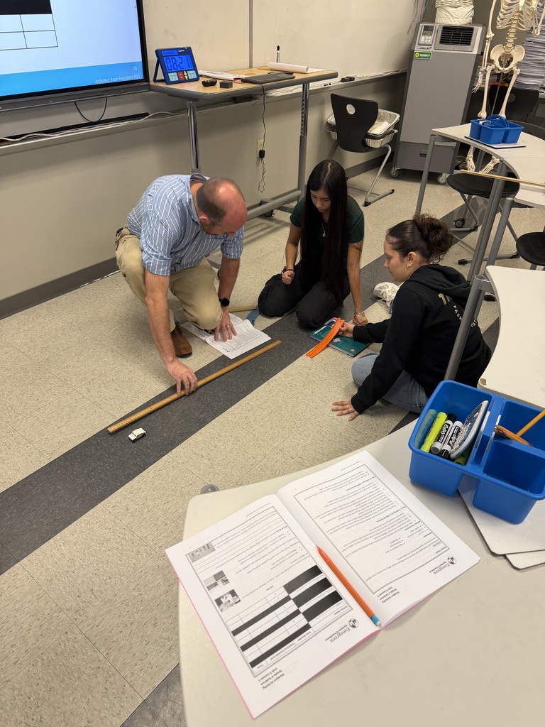 Three people are working on the floor in a classroom. A man bends over papers, a woman kneels with a pencil, and another woman sits. Equipment and documents are on tables.