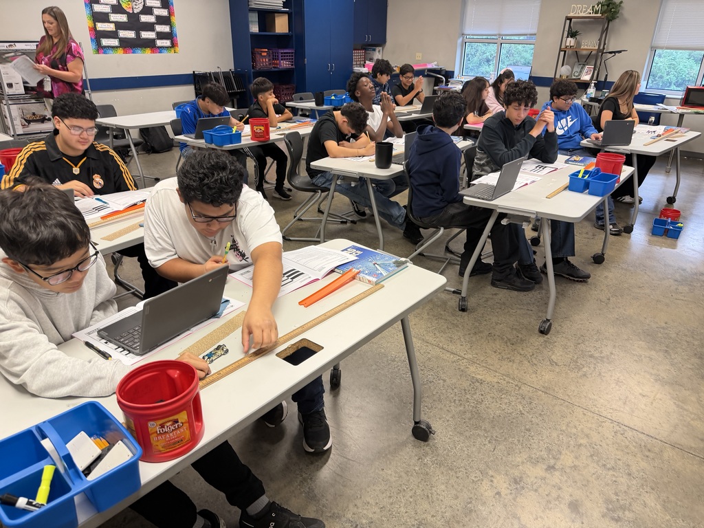 Students sit at tables in a classroom, engaged in various activities. They have laptops and tools on their desks.