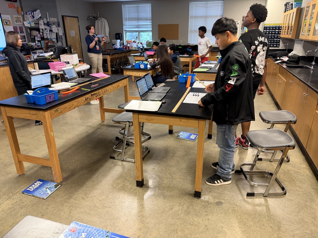 Students work on laptops and papers at tables in a room with a concrete floor and cabinets.