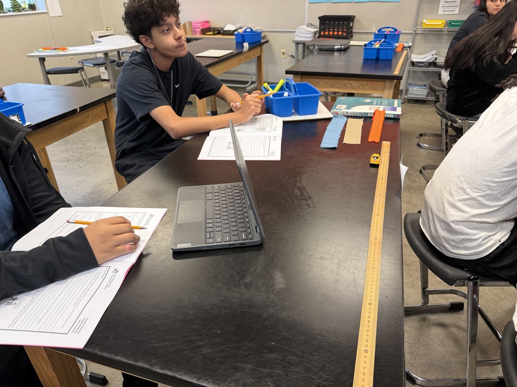 Students seated at tables in a classroom, working on papers with a laptop and ruler in front.