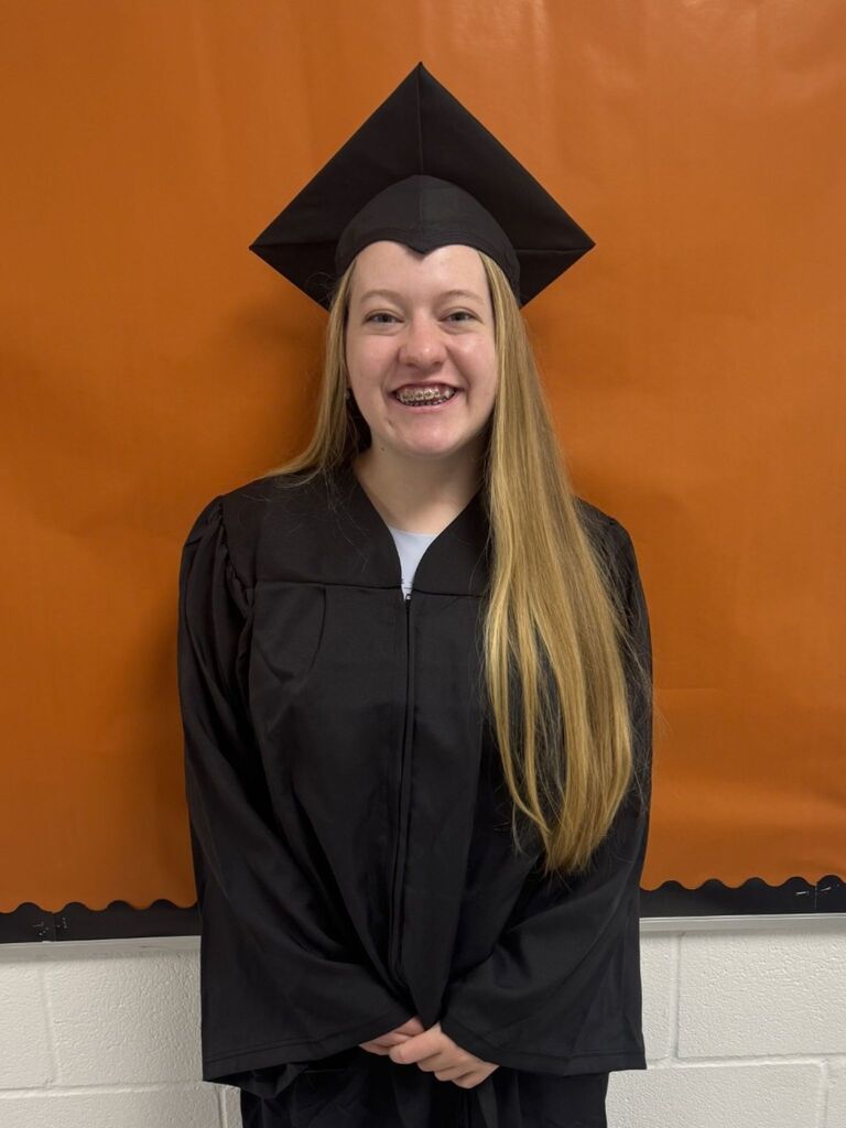Woman in a black graduation gown and cap, smiling at the camera against an orange backdrop.