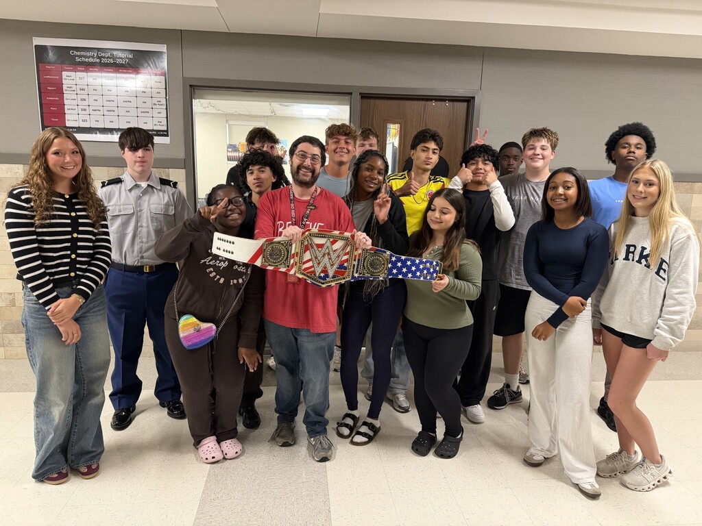 A group of students and staff posing in a hallway with a championship belt held by a student.