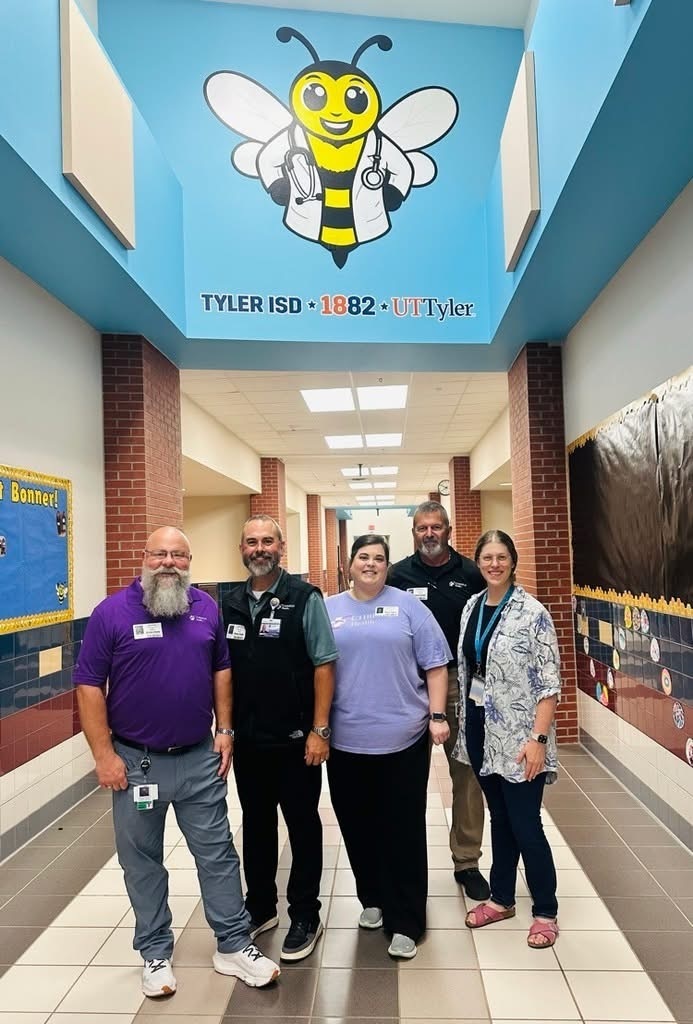 Five people stand in a hallway with a bee logo above. They wear different colored tops and pants.
