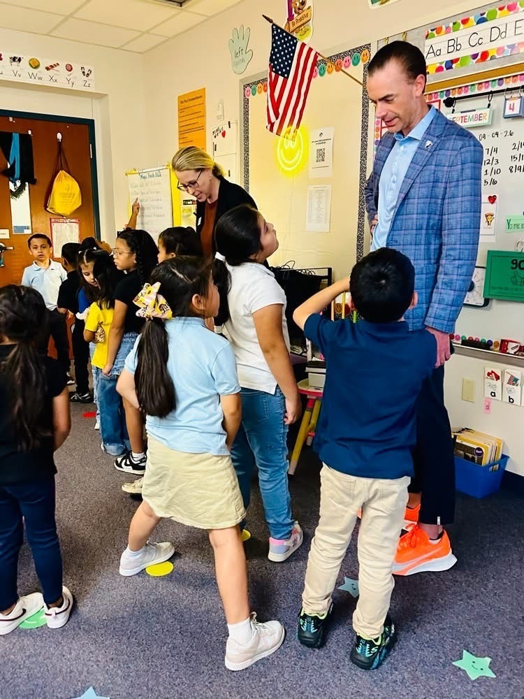 A man and woman standing in a classroom with children. The man wears a blue suit, and the woman wears glasses.