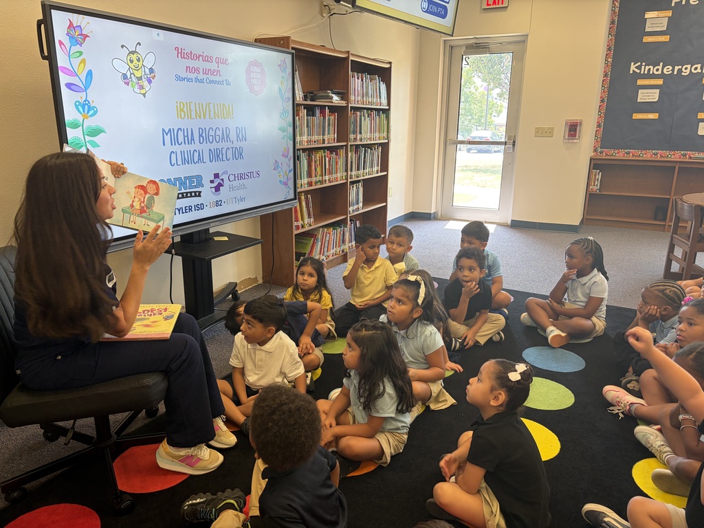 A woman reads a book to young children in a classroom, with bookshelves in the background.