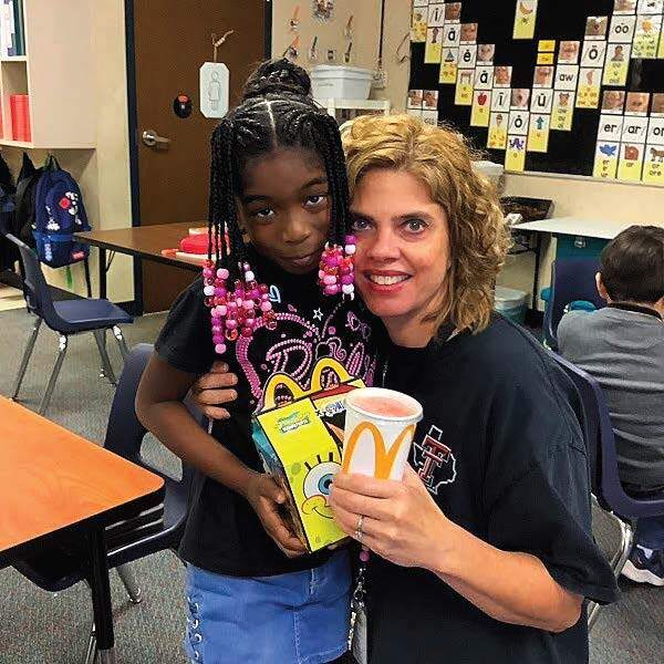 A woman and a girl stand in a classroom. The girl holds a box and cup. They smile and pose for a photo.