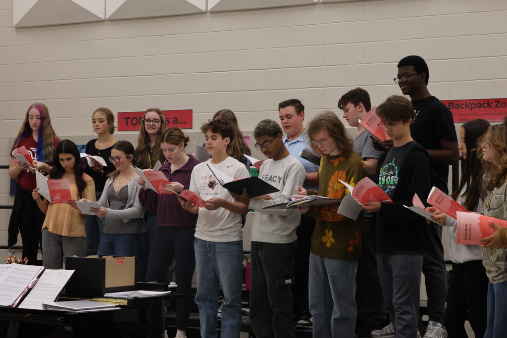 A group of students, some holding music sheets, stand in a room with a piano and red banners in the background.