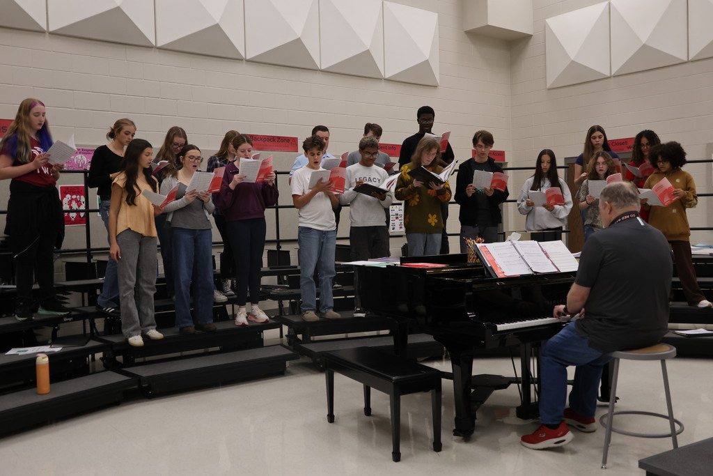 A choir rehearses in a room with a piano, facing the conductor. The room has bleachers, a white ceiling, and a white wall.