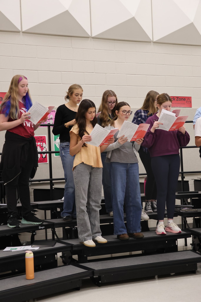 Group of people on raised platforms in a gym, holding papers, and appearing to rehearse.