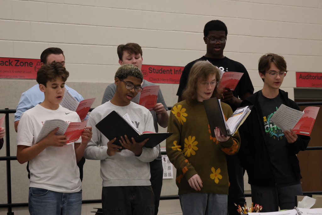 A group of students hold red songbooks, some wearing glasses, in a room with a white wall.