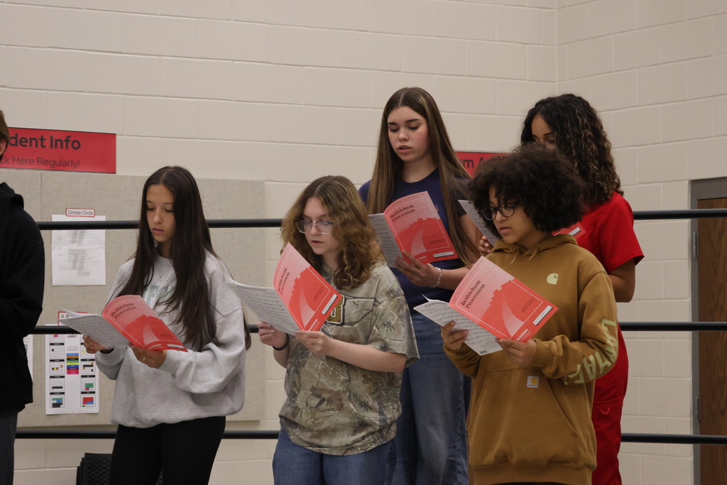 Six people, possibly students, holding red pamphlets, stand in a classroom. A man in black stands to the left.