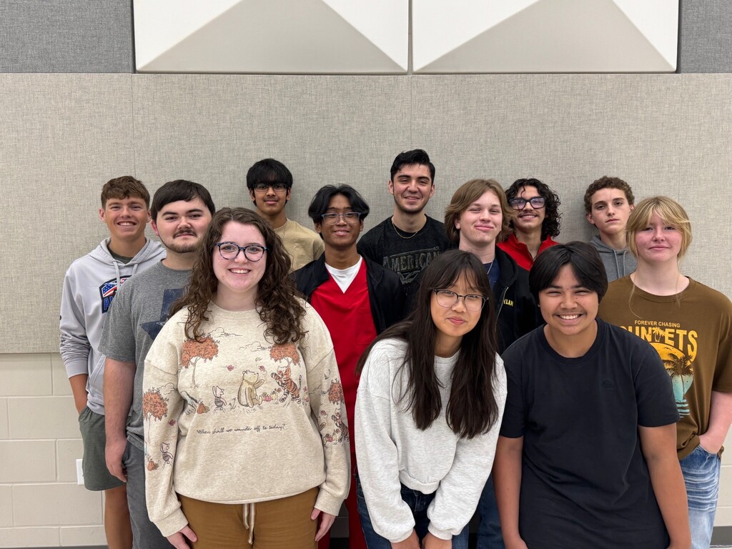 A group of students stand together inside a room, with a wall pattern behind them.