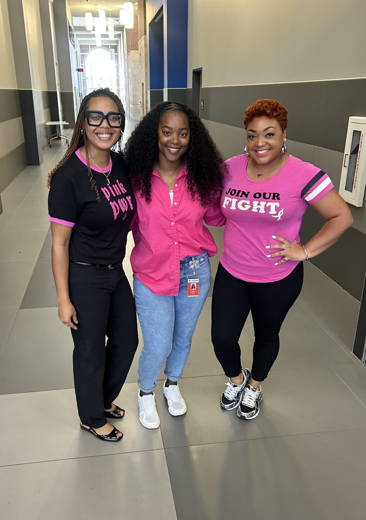 Three women in pink shirts pose together in a brightly lit hallway. The middle woman wears a pink top.