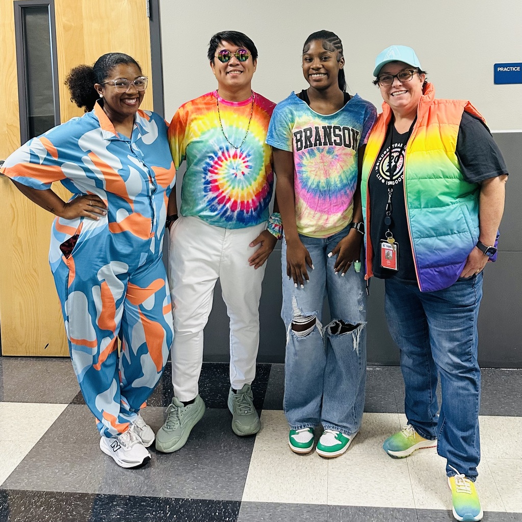 Four women in matching outfits, with one wearing a rainbow vest, pose for a photo indoors.