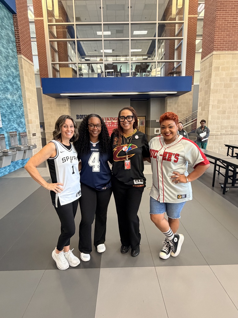 Four individuals stand in a hallway, smiling and posing. One wears a blue shirt with number 4, another has a black shirt.