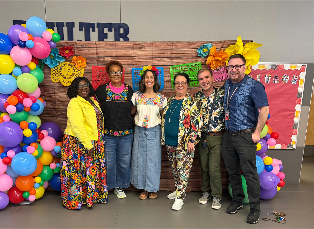 Six adults in front of a wooden backdrop, colorful balloons, and decorations. They smile for a photograph.