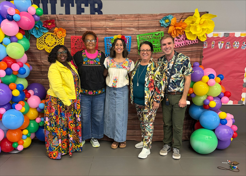Five adults pose together in front of a colorful backdrop with vibrant balloons and decorations.