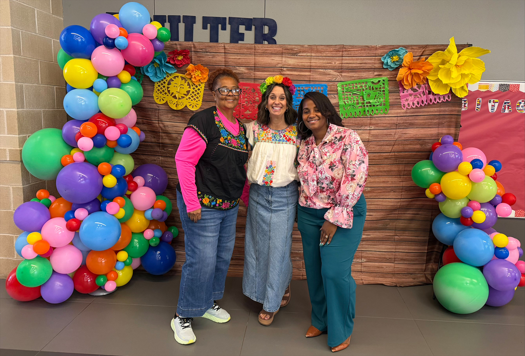Three women pose together in front of a backdrop of colorful balloons and a wooden panel.