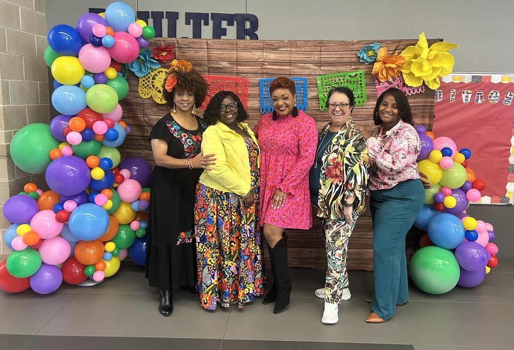 Five women in various outfits standing by a colorful balloon display and a wooden banner.