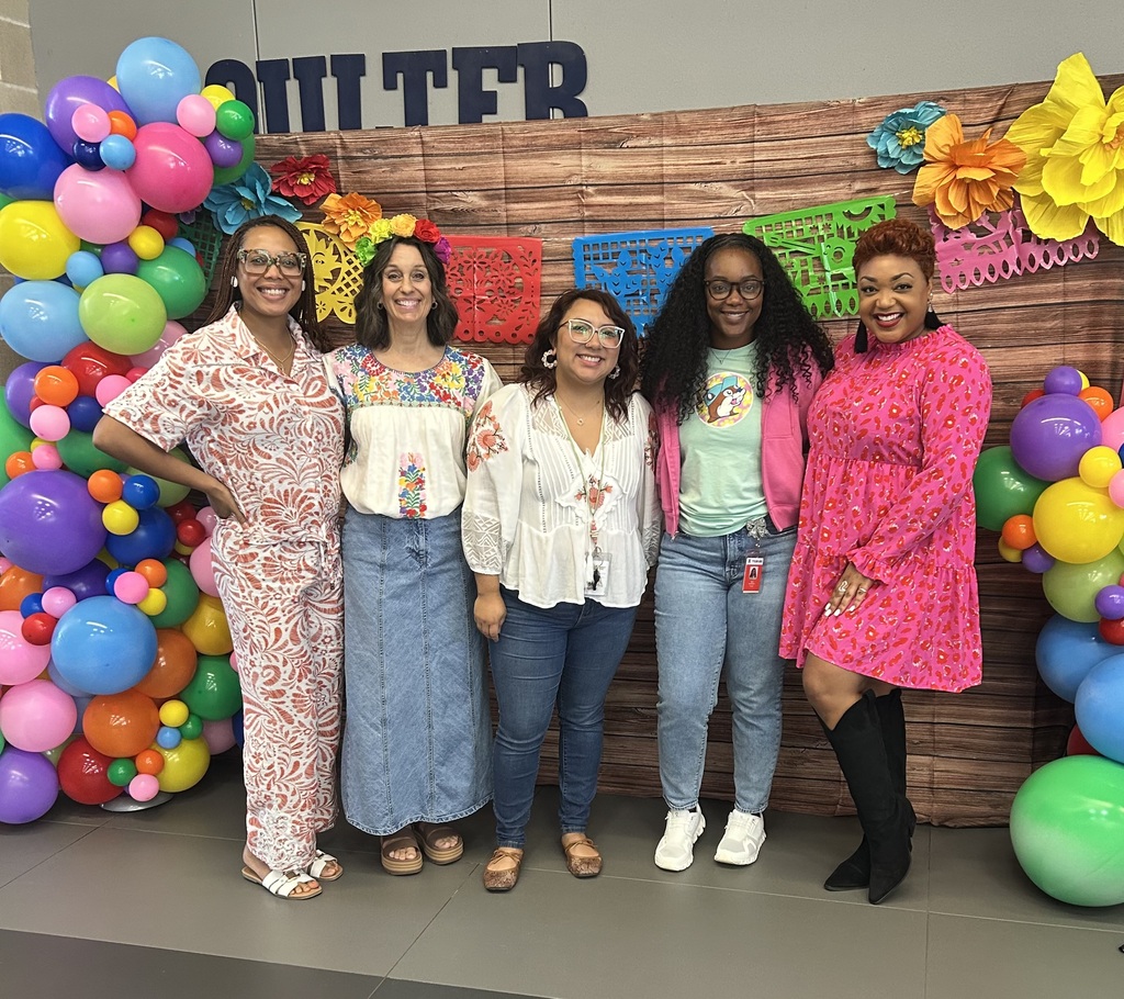 Six women stand before a wooden backdrop with a colorful banner and balloons. The backdrop is decorated with multicolored flowers.