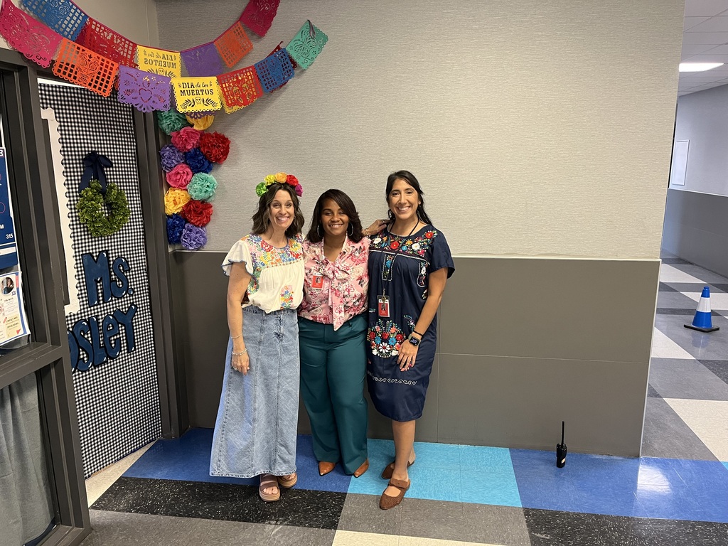 Three women in colorful dresses stand next to each other in a hallway with a white wall and blue carpet.